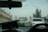 The scene is from inside a vehicle, looking through the front windshield at traffic on a bridge. Cars are lined up, with a green and yellow taxi in front. The bridge structure is visible ahead with towers and overhead traffic lights. To the right, there is a large tree and to the left, a row of buildings with satellite dishes and utility wires.