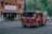 A red fire truck is driving through a city street. The truck has visible markings indicating it is part of an emergency service. Next to the street is an older-style building with a theater marquee displaying advertisements for a comedy play. The area is surrounded by other urban buildings, trees, and parked cars.
