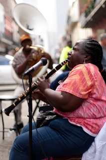 A street musician deeply engaged in playing a clarinet with a microphone in front of her, surrounded by an urban setting. In the background, another musician with a large brass instrument can be seen, and the street is lined with buildings.