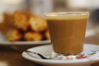 A glass of coffee with foamy top served on a white saucer, accompanied by a spoon. In the background, there are blurred golden-brown pastries on a white plate.