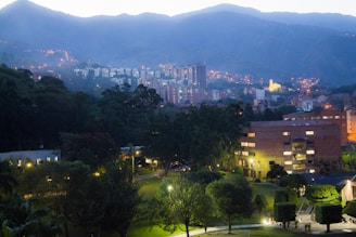 A cityscape with buildings nestled at the base of hills, illuminated with streetlights and building lights against the backdrop of misty mountains. Lush greenery and a well-lit park with trees are visible in the foreground. The atmosphere suggests an urban area surrounded by nature, blending urban development with natural elements.