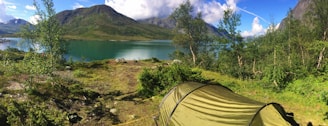 A serene camping scene by a tranquil lake surrounded by lush greenery and mountains in the background. The green tent is prominently placed in the foreground amidst the natural landscape. The sky is clear with a few scattered clouds, adding to the peaceful ambiance.