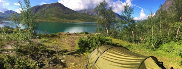 A serene camping scene by a tranquil lake surrounded by lush greenery and mountains in the background. The green tent is prominently placed in the foreground amidst the natural landscape. The sky is clear with a few scattered clouds, adding to the peaceful ambiance.