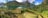 A serene camping scene by a tranquil lake surrounded by lush greenery and mountains in the background. The green tent is prominently placed in the foreground amidst the natural landscape. The sky is clear with a few scattered clouds, adding to the peaceful ambiance.