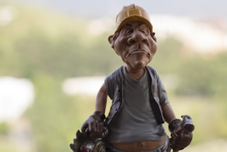A caricature sculpture of a worker wearing a yellow hard hat and casual clothing. The figure has exaggerated facial features and holds tools in both hands. The background is blurred and suggests an outdoor setting with greenery.