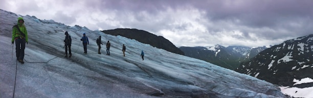 A group of people are trekking across a vast, icy glacier with snow-capped mountains in the background under a cloudy sky. They are connected by a rope for safety, wearing snow gear and carrying climbing equipment. The scene conveys a sense of adventure and exploration amid a rugged natural landscape.