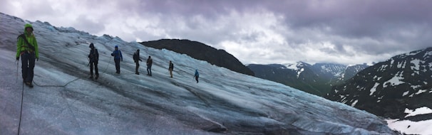 A group of people are trekking across a vast, icy glacier with snow-capped mountains in the background under a cloudy sky. They are connected by a rope for safety, wearing snow gear and carrying climbing equipment. The scene conveys a sense of adventure and exploration amid a rugged natural landscape.