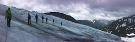 A group of people are trekking across a vast, icy glacier with snow-capped mountains in the background under a cloudy sky. They are connected by a rope for safety, wearing snow gear and carrying climbing equipment. The scene conveys a sense of adventure and exploration amid a rugged natural landscape.