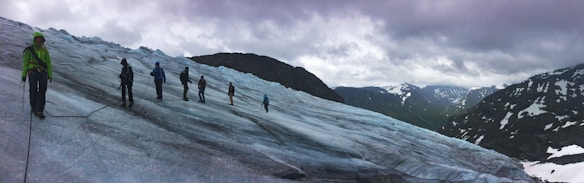 A group of people are trekking across a vast, icy glacier with snow-capped mountains in the background under a cloudy sky. They are connected by a rope for safety, wearing snow gear and carrying climbing equipment. The scene conveys a sense of adventure and exploration amid a rugged natural landscape.