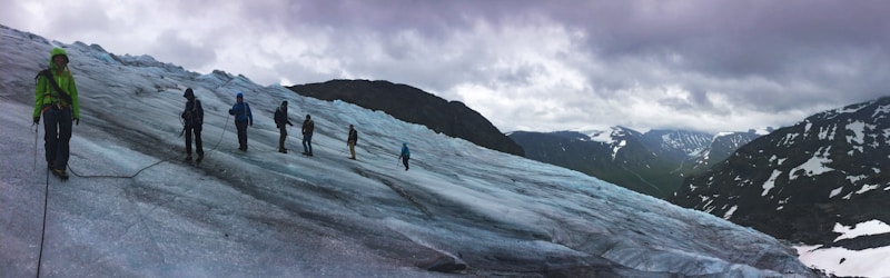 A group of people are trekking across a vast, icy glacier with snow-capped mountains in the background under a cloudy sky. They are connected by a rope for safety, wearing snow gear and carrying climbing equipment. The scene conveys a sense of adventure and exploration amid a rugged natural landscape.