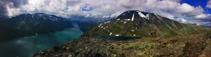 Panoramic view of a stunning mountainous landscape with a clear turquoise lake nestled between towering snow-capped peaks. A group of hikers stands on the rocky foreground, admiring the vast, serene natural beauty surrounding them. The sky is partly cloudy, casting varied shadows and light across the scene.