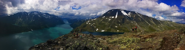Panoramic view of a stunning mountainous landscape with a clear turquoise lake nestled between towering snow-capped peaks. A group of hikers stands on the rocky foreground, admiring the vast, serene natural beauty surrounding them. The sky is partly cloudy, casting varied shadows and light across the scene.