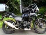 A parked Royal Enfield Himalayan motorcycle is positioned on a road with a lush green forest backdrop. The bike has saddlebags and a box attached to it, suggesting it is equipped for travel. A small waterfall is visible in the background, giving a sense of a tranquil outdoor setting.