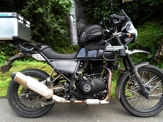 A parked Royal Enfield Himalayan motorcycle is positioned on a road with a lush green forest backdrop. The bike has saddlebags and a box attached to it, suggesting it is equipped for travel. A small waterfall is visible in the background, giving a sense of a tranquil outdoor setting.