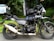 A parked Royal Enfield Himalayan motorcycle is positioned on a road with a lush green forest backdrop. The bike has saddlebags and a box attached to it, suggesting it is equipped for travel. A small waterfall is visible in the background, giving a sense of a tranquil outdoor setting.