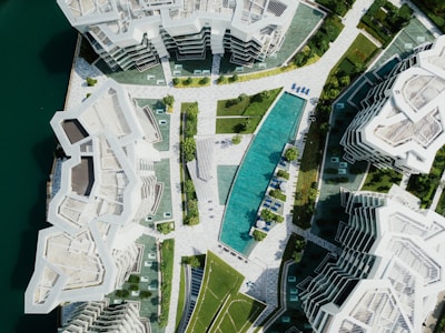 Aerial view of modern, geometric residential buildings surrounded by greenery. A long, narrow pool is centrally located, flanked by lounge chairs. The architecture features angular designs with white facades and rooftop terraces.