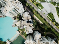 An aerial view of a modern residential complex featuring multiple high-rise buildings with light-colored roofs and structured layouts. The buildings are surrounded by lush greenery and a large swimming pool with turquoise water. Adjacent to the complex is a neatly maintained road with a bus and landscaped gardens. The overall setting is urban with a touch of natural elements.