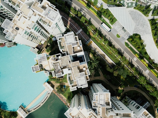 An aerial view of a modern residential complex featuring multiple high-rise buildings with light-colored roofs and structured layouts. The buildings are surrounded by lush greenery and a large swimming pool with turquoise water. Adjacent to the complex is a neatly maintained road with a bus and landscaped gardens. The overall setting is urban with a touch of natural elements.