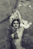 A woman floats on her back in the water, wearing a polka dot bikini. Her arms are extended above her head, and her eyes are closed. The image is captured in a monochrome tone, giving it a vintage feel. Light reflects off the water, creating a serene atmosphere.