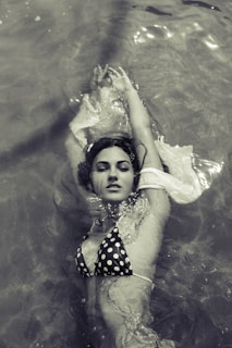 A woman floats on her back in the water, wearing a polka dot bikini. Her arms are extended above her head, and her eyes are closed. The image is captured in a monochrome tone, giving it a vintage feel. Light reflects off the water, creating a serene atmosphere.
