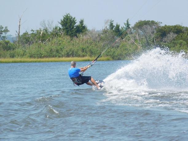 A person wearing a blue shirt is engaged in kiteboarding or kite surfing on a body of water. The individual is leaning back, holding onto the control bar attached to the kite, which is not visible. The water is disturbed by their movements, creating a trail of spray and ripples. Surrounding the water is a lush green backdrop with trees and shrubs visible in the distance.