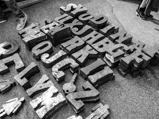 Large metal letters and shapes scattered on the ground, arranged in a somewhat random pattern. The letters are three-dimensional and cast shadows on the concrete surface. Two pairs of shoes are visible at the edges, suggesting people are nearby or passing through.