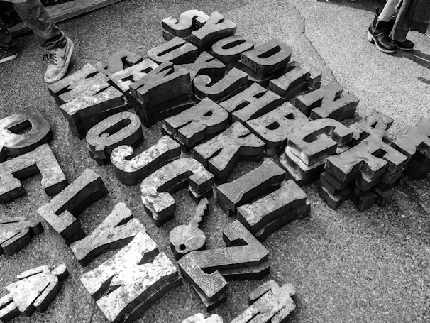 Large metal letters and shapes scattered on the ground, arranged in a somewhat random pattern. The letters are three-dimensional and cast shadows on the concrete surface. Two pairs of shoes are visible at the edges, suggesting people are nearby or passing through.
