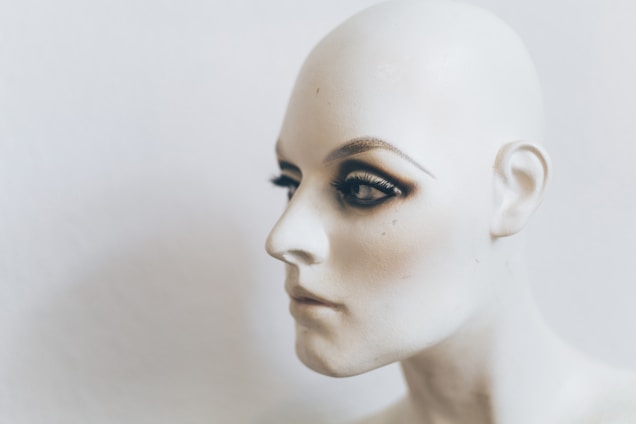 A close-up profile of a mannequin head featuring lifelike facial details and makeup, including prominently applied eyeshadow and false eyelashes. The head is bald, and the skin tone is a smooth, pale color against a neutral background.