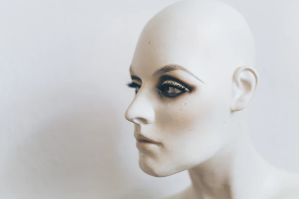 A close-up profile of a mannequin head featuring lifelike facial details and makeup, including prominently applied eyeshadow and false eyelashes. The head is bald, and the skin tone is a smooth, pale color against a neutral background.