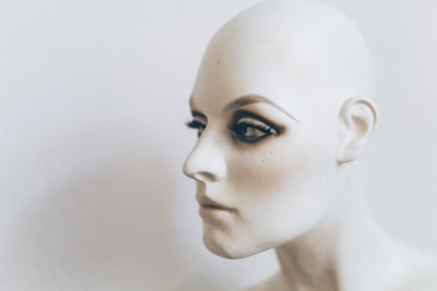 A close-up profile of a mannequin head featuring lifelike facial details and makeup, including prominently applied eyeshadow and false eyelashes. The head is bald, and the skin tone is a smooth, pale color against a neutral background.