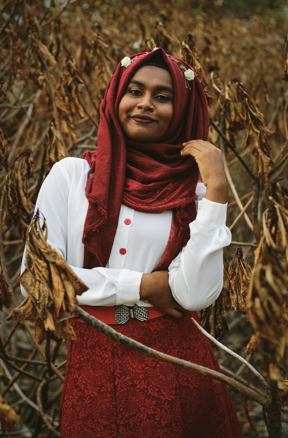 femme portant un hijab rouge souriant derrière des feuilles séchées brunes