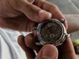 A person is holding a watch mechanism in their hands, revealing the intricate gears and components of the watch's inner workings. The image focuses on the metal parts and craftsmanship of the watch movement.