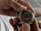 A person is holding a watch mechanism in their hands, revealing the intricate gears and components of the watch's inner workings. The image focuses on the metal parts and craftsmanship of the watch movement.