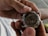 A person is holding a watch mechanism in their hands, revealing the intricate gears and components of the watch's inner workings. The image focuses on the metal parts and craftsmanship of the watch movement.