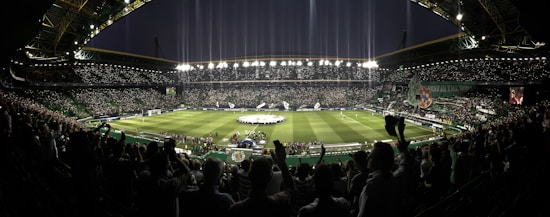 A large, packed stadium under night lights with a vast crowd surrounding the pitch, where a group of people appear to be performing or gathering in a ceremonial setup. Bright floodlights illuminate the scene, and a large screen displays an image. The atmosphere is filled with excitement and anticipation, with many fans captured in the foreground waving and cheering.