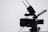 A professional video camera with a large lens and an attached external monitor. Two antennas are mounted on top, likely part of a wireless transmission system. The camera is on a tripod, set against a simple, light-colored background.