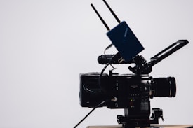 A professional video camera with a large lens and an attached external monitor. Two antennas are mounted on top, likely part of a wireless transmission system. The camera is on a tripod, set against a simple, light-colored background.