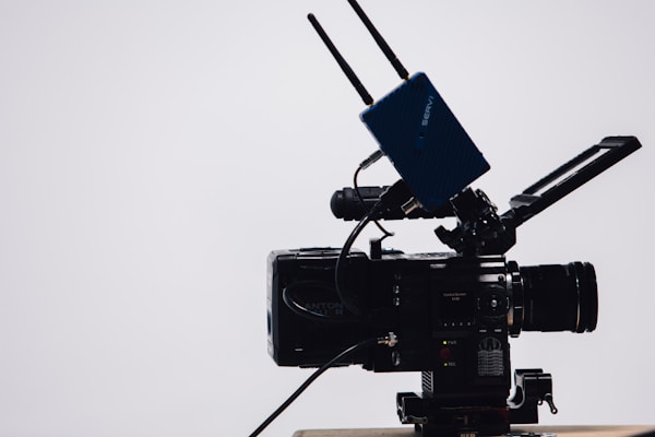 A professional video camera with a large lens and an attached external monitor. Two antennas are mounted on top, likely part of a wireless transmission system. The camera is on a tripod, set against a simple, light-colored background.