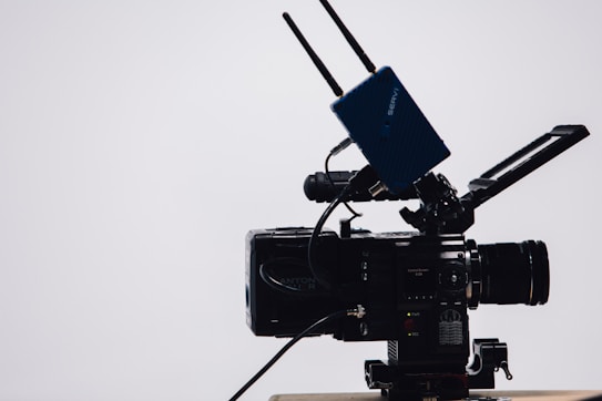 A professional video camera with a large lens and an attached external monitor. Two antennas are mounted on top, likely part of a wireless transmission system. The camera is on a tripod, set against a simple, light-colored background.