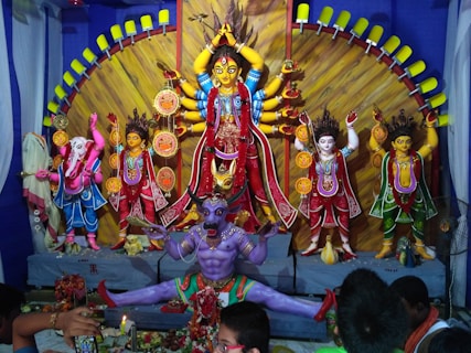 The image displays a vibrant and intricately decorated religious scene with various Hindu deities. The central figure is a multi-armed goddess dressed in red and adorned with jewelry, surrounded by other deity statues including the elephant-headed god, Ganesha. A fierce demon in purple is positioned at the foreground depicting a powerful scene of divinity over evil.