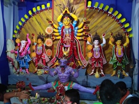 The image displays a vibrant and intricately decorated religious scene with various Hindu deities. The central figure is a multi-armed goddess dressed in red and adorned with jewelry, surrounded by other deity statues including the elephant-headed god, Ganesha. A fierce demon in purple is positioned at the foreground depicting a powerful scene of divinity over evil.