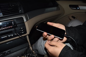 A person is holding a smartphone plugged into a car's audio system. The display on the audio system shows the word 'STOP', and multiple cables are visible nearby. The interior of the car features beige and black elements, including the dashboard and seat covers.