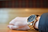 A close-up of a wrist wearing a luxurious gold watch with a detailed black dial and intricate design. The background is softly blurred, highlighting the watch and wrist.
