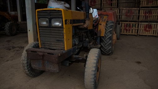 A worn, rustic tractor is parked inside a dimly lit barn. The tractor shows signs of use, with faded yellow and orange paint and visible rust. Its large tires are covered in dust, and behind it are stacks of wooden crates in the background.