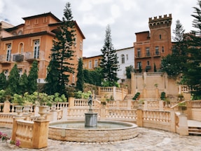 A picturesque courtyard with elegant Mediterranean-style architecture, featuring warm-colored buildings with arched windows and ornate balconies. A tiered garden with various green plants and trees enhances the scenery. In the foreground, a circular stone fountain is adorned with a sculpted figure.