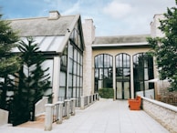 A well-maintained building with a modern architectural design is surrounded by lush green trees. It features large arched windows with reflective glass and a slanted roof structure. The exterior is made of light-colored stone, complemented by a spacious paved pathway leading to the entrance. A vibrant orange bench and a neatly trimmed hedge add to the aesthetic appeal.