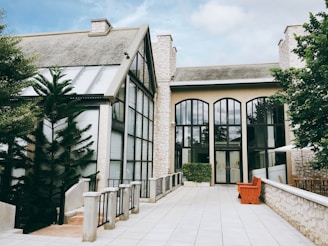 A well-maintained building with a modern architectural design is surrounded by lush green trees. It features large arched windows with reflective glass and a slanted roof structure. The exterior is made of light-colored stone, complemented by a spacious paved pathway leading to the entrance. A vibrant orange bench and a neatly trimmed hedge add to the aesthetic appeal.