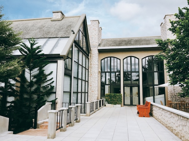 A well-maintained building with a modern architectural design is surrounded by lush green trees. It features large arched windows with reflective glass and a slanted roof structure. The exterior is made of light-colored stone, complemented by a spacious paved pathway leading to the entrance. A vibrant orange bench and a neatly trimmed hedge add to the aesthetic appeal.