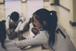 A group of athletes wearing sports uniforms is seated on the floor in a gymnasium, engaging in stretching exercises. The focus is on one athlete with a ponytail, wearing a jersey with the number seven, smiling as she stretches her legs. The lighting is soft, creating a relaxed atmosphere.