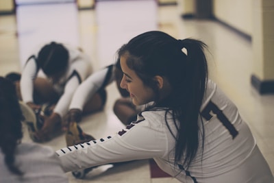 A group of athletes wearing sports uniforms is seated on the floor in a gymnasium, engaging in stretching exercises. The focus is on one athlete with a ponytail, wearing a jersey with the number seven, smiling as she stretches her legs. The lighting is soft, creating a relaxed atmosphere.